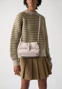Borsa a tracolla beige con design a patta strato e accenti cuciti. Indossata con un maglione a righe e una gonna plissettata verde oliva.