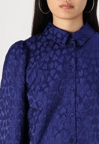 Purple blouse featuring a textured pattern of small hearts, a collar, and front buttons. The fabric exhibits a smooth, shiny finish.