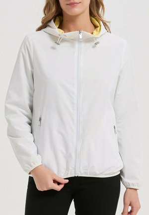 Femme portant une veste à capuche blanche zippée avec une doublure jaune, un pantalon noir, tenant l'ourlet inférieur de la veste.