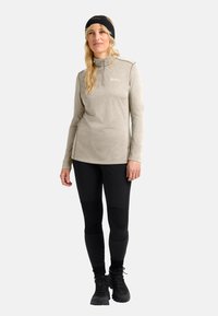 Donna in felpa beige con zip, leggings neri, scarponi da trekking neri e fascia nera per capelli che si staglia su uno sfondo semplice.