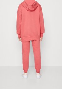 En person iförd en rosa hoodie och matchande sweatpants, med vita sneakers, vänd bort från kameran mot en enkel vit bakgrund.