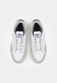 Zapatillas blancas con parte superior de piel, acentos grises, detalles perforados y un logo azul de Adidas en la lengüeta y el costado.