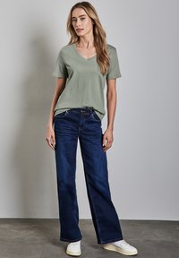 Světle zelené tričko s výst�řihem do V a krátkými rukávy zkombinované s tmavě modrými širokými džíny a bílými teniskami, s neutrálním pozadím.