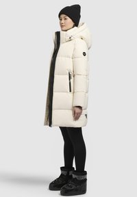 Manteau rembourré crème avec une silhouette ajustée, accents de fermeture éclair noire et grande capuche ; associé à des leggings noirs et des bottes isolées.