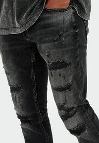 Jean noir déchiré avec des accents décolorés, présentant plusieurs déchirures et des bords effilochés, en matériau en denim, design slim fit.