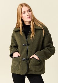 Giacca in pile verde oliva con dettagli neri, due tasche anteriori e un ampio colletto; presenta bottoni a cordino e una texture morbida.
