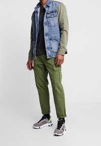 Farkkutakki oliivinvihreillä hihoilla, harmaa t-paita, vihreät cargo-housut ja harmaat tennarit mustilla ja neonkorostuksilla. Sisältää taskuja.