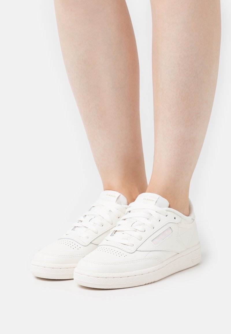 Des baskets blanches présentent un dessus en cuir lisse, un bout perforé, des lacets ronds et une semelle en caoutchouc texturée ; accents de couleur mineurs près des côtés.