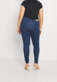 Kvinna som bär mörkblå skinny jeans och nakna blockklackssandaler, stående mot en enkel ljus bakgrund, vänd bort.