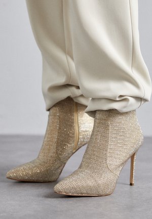 Bottines beige à talons hauts avec des strass scintillants, portées avec un pantalon ample de couleur crème sur un sol gris.