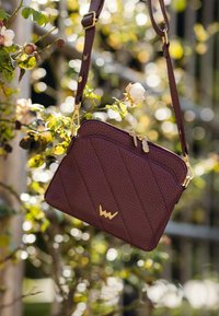 Texturovaná burgundská crossbody taška s prošívaným designem, zlatými kovovými prvky a zipovým uzávěrem. Na přední straně má logo ve zlaté barvě.