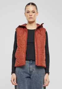 Gestepptes, rostfarbenes Weste mit Kapuze, Frontreißverschluss und seitlichen Taschen, getragen über einem schwarzen Oberteil und kombiniert mit lockeren Jeans.