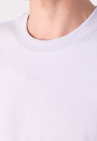 Zbliżenie na osobę w jasnofioletowej koszulce z okrągłym dekoltem, na której piersi znajduje się wytłoczone logo "HUGO".