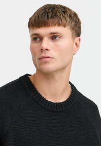 Pulover negru tricotat cu guler rotund cu nervuri și mâneci raglan, având un finisaj texturat. Modelul are părul scurt de culoare brunetă și ochi albaștri.