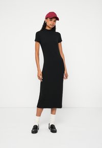 Melna, īsām piedurknēm, pieguloša kleita ar apaļu kakla izgriezumu, kas sniedzas līdz pusstilbam. Valkāta ar baltiem, teksturētiem zeķēm un melniem loaferiem, papildināta ar tumši sarkanu cepuri.