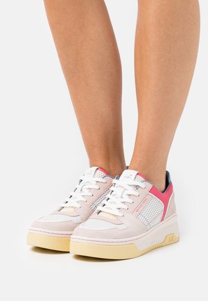 Paire de baskets à plateforme roses et blanches avec des lacets blancs portées sur des jambes nues contre un fond uni.