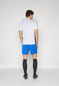 Witte korte mouwen shirt met zwarte accenten, in combinatie met blauwe shorts en zwarte kniekousen, gedragen met zwarte voetbalschoenen.
