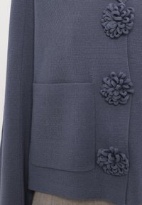 Kötött sötétkék kardigán, amely texturált anyagból készült, elülső zsebe van, és három szövet virág alakú díszgombbal rendelkezik.