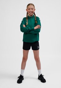 Grøn langærmet pullover med halv lynlås, kombineret med sorte shorts. Har hvidt mærke, fletninger som frisure og sorte sportssko.