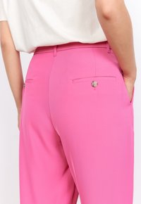 Un pantalon rose avec une texture lisse, doté de deux poches arrière et d'accents en bouton. Le design comprend une coupe ajustée et une taille mi-haute.