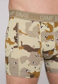Camo-patterned boxer shorts in beige, brown, and tan with a soft elastic waistband labeled "CAMP DAVID." Features visible stitching and fit.