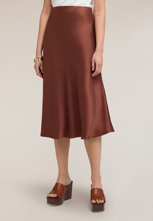 Bruine satijnen midi-rok gedragen met bruine platform mule sandalen en gouden armbanden om de pols, getoond op het onderste deel van het lichaam tegen een neutrale achtergrond.