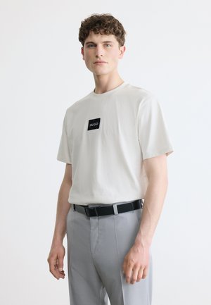 Mladý muž s krátkymi kučeravými vlasmi, oblečený v bielom tričku s logom Hugo, svetlosivých nohaviciach a čiernom opasku, stojaci pred jednoduchým pozadím.