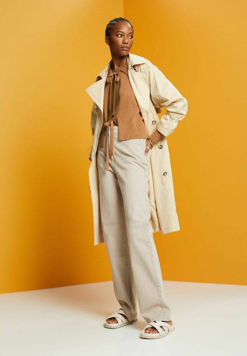 Manteau trench beige clair, pull marron à nouer à l'avant, pantalon clair et sandales blanches à lanières. Textures lisses et palette de couleurs neutres.