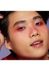 Närbild av ett ansikte med färgglad make-up: gradient lila till röd ögonskugga, glittrande detaljer på kinder och näsa, ljus hudton och glansiga läppar.