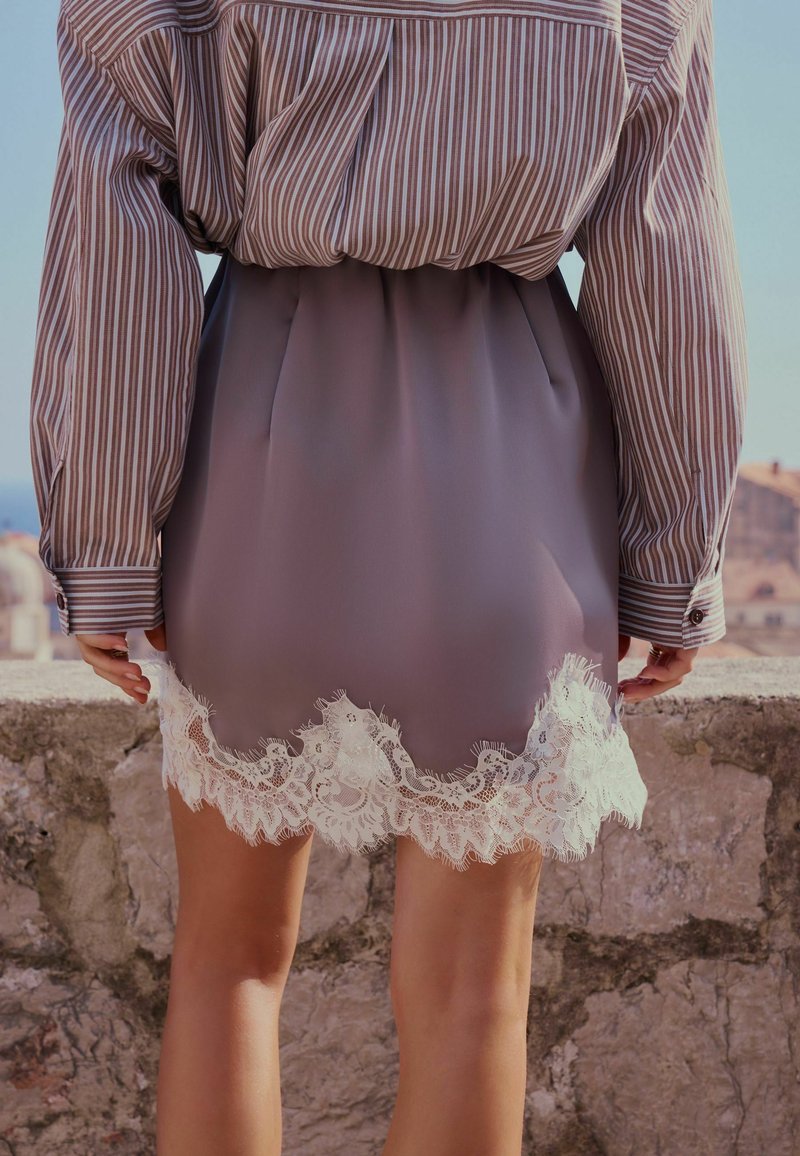 Person wearing a purple skirt with white lace trim and a tucked-in striped long-sleeve shirt, standing by a stone wall outdoors.