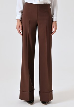 Pantalons marron à jambes larges en tissu lisse, avec une taille haute, des plis à l'avant et des poignets repliés. Portés avec un chemisier blanc et des chaussures noires.
