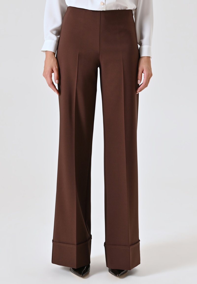 Pantalons marron à jambes larges en tissu lisse, avec une taille haute, des plis à l'avant et des poignets repliés. Portés avec un chemisier blanc et des chaussures noires.
