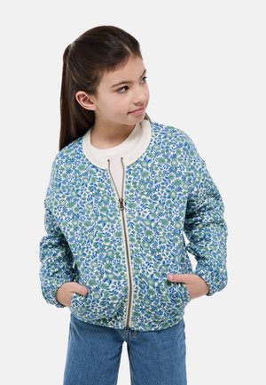 Fille aux longs cheveux bruns portant une veste zippée fleurie bleue et verte et un jean bleu, les mains dans les poches, regardant sur le côté.