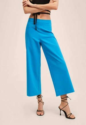 Vrouw draagt felblauwe wijde cropped broek met zwarte sandalen met hoge hak en bandjes, en een zwart cropped topje met gestrikte bandjes.