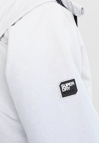 Lys grå hættetrøje med en glat tekstur, der har et sort "SUPER DRY" logo patch på ærmet. Enkel, moderne design, ingen synlige mønstre.