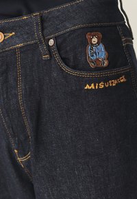 Sötét farmernadrág narancssárga varrással és egy teddy maci folttal az elülső zseben. Sárgával hímzett "MISUSZCZEK" felirat az oldalon.