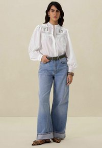 Chemise blanche à manches bouffantes et détails en dentelle, associée à un jean large bleu clair et une ceinture verte ; chaussures à imprimé léopard.