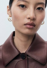 Brown leather jacket with a large collar, fastened by a black button. Model features hoop earrings and smooth skin with neutral tones.