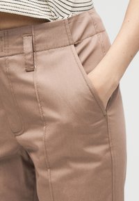 Άτομο που φοράει μπεζ παντελόνια με το χέρι στην μπροστινή τσέπη και μια ριγέ μπλούζα μερικώς ορατή στη μέση.