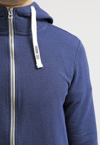 Felpa azul marino con cremallera completa en la parte delantera, tejido texturizado, cordones ajustables blancos y etiqueta de marca en el cordón derecho.