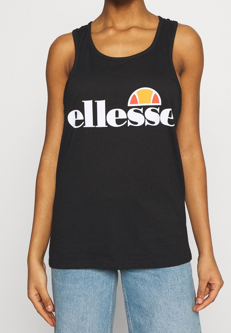 Person, die ein schwarzes ärmelloses Oberteil mit weißem "ellesse"-Text und Logo trägt, kombiniert mit hellblauen Jeans, steht vor einem einfarbigen Hintergrund.