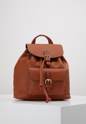 Brązowy skórzany plecak z przednią kieszenią, złotymi klamrami, zapięciem na sznurek i górnym uchwytem, na białej powierzchni ekspozycyjnej.