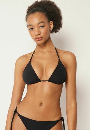 Mujer con cabello rizado que lleva un bikini negro de triángulo con tirantes halter, de pie contra un fondo claro y liso.