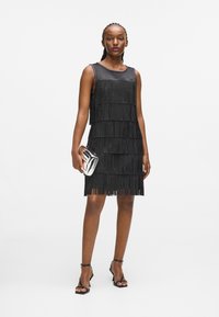 Fekete ujj nélküli ruha réteges bojtokkal, puha anyagból készült. A modellen ezüst színű retikült tart, fekete pántos magassarkúval párosítva.