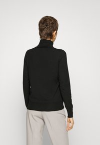 Pull à col roulé noir en matériau doux, présentant un design ajusté, des poignets et un ourlet côtelés, vu de dos avec un pantalon clair.