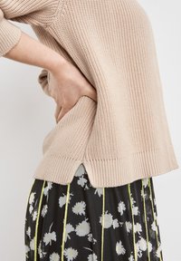 Beige pletený svetr s žebrovanými manžetami a lemem, se bočními rozparky. V kombinaci s černou sukní zdobenou bílým květinovým vzorem a žlutými akcenty.