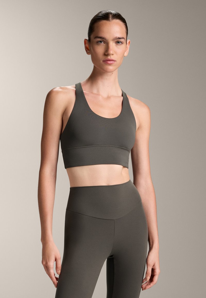Oljčno zelena športna modrček z zaobljenim izrezom in širokimi naramnicami. V kombinaciji z visokim pasom in oprijetimi legginsi, oboje iz gladkega materiala.