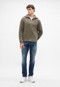Pullover a coste color khaki con colletto a mezza zip, indossato sopra una camicia bianca, abbinato a jeans blu e sneakers bianche.