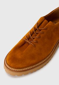 Des chaussures en daim d'une riche teinte orange-brune, présentant un bout rond, cinq œillets et une semelle en caoutchouc beige robuste avec des détails de couture.
