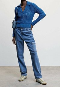 Blau gestrickter Pullover mit V-Ausschnitt und Kragen, kombiniert mit hellblauen Cargo-Jeans und beigefarbenen Sneakers, dazu eine kleine Handtasche.